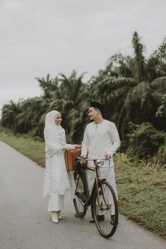 a man and woman standing next to a bike on a road with palm trees in the background