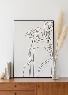 a vase with some flowers on top of a wooden dresser next to a framed art piece