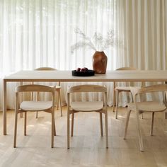 a dining table with chairs and a vase on the top, in front of a curtained window