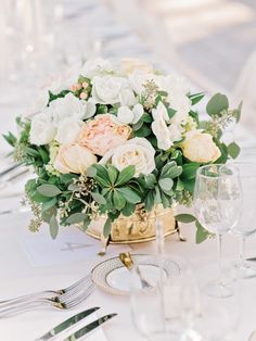 an arrangement of flowers and greenery sits on a table set for a formal dinner