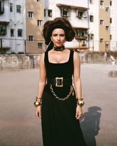 a woman in a black dress and gold jewelry standing on the street with buildings behind her