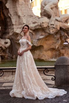 a woman standing in front of a fountain wearing an off the shoulder wedding dress with flowers on it