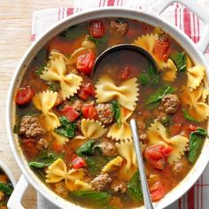 a bowl of pasta soup with meat, spinach and tomatoes on the side next to a spoon