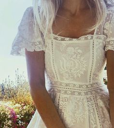 a woman in a white dress is standing near some flowers and holding her hand on her hip