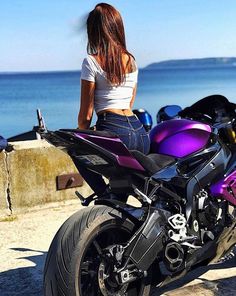 a woman sitting on top of a purple motorcycle next to the ocean and looking at the water