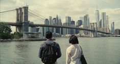 two people looking out over the water in front of a large bridge and cityscape