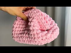 a hand holding a pink crochet purse over a white wall with curtains in the background