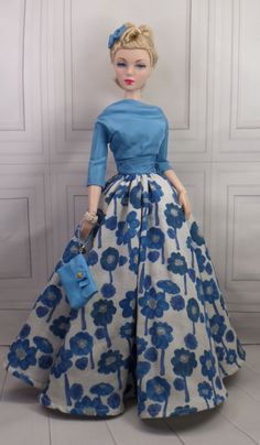 a doll wearing a blue and white dress with flowers on it's skirt, holding a purse