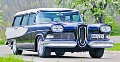 an old black and white station wagon driving down the road with trees in the background