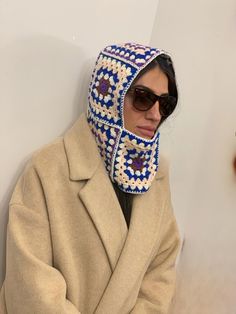 a woman wearing a crocheted hat and coat with sunglasses on her head is leaning against a wall