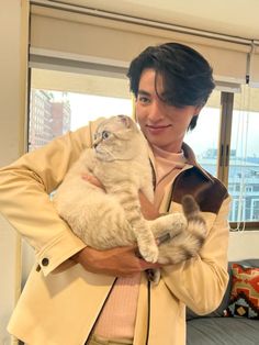 a man holding a cat in his arms while standing next to a couch and window