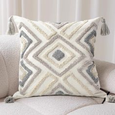 a white and grey pillow with tassels sitting on top of a couch next to a window