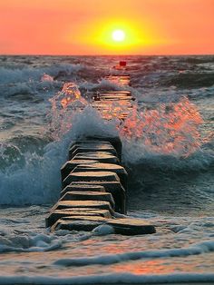 the sun is setting over the ocean with waves crashing on it and a long pier in the foreground