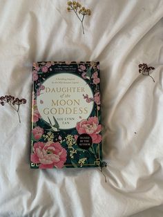 the book daughter of the moon goddess is laying on a bed with white sheets and pink flowers