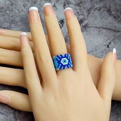 Protect yourself in style with this stunning Evil Eye beaded ring, expertly handcrafted from high-quality Delica beads. This wide ring showcases a captivating design in shades of blue, light blue, white, and silver, symbolizing protection and good fortune. The intricate pattern and bold colors make it a unique and eye-catching accessory, perfect for adding a touch of mystique to any outfit. Ideal for those who appreciate unique, artisan jewelry, this ring is perfect for everyday wear or as a tho Blue Beaded Rings As A Gift, Evil Eye Beaded Ring, Spiritual Blue Rings With Evil Eye Detail, Adjustable Blue Evil Eye Bracelet Hand-strung, Adjustable Blue Hand-strung Evil Eye Bracelet, Artisan Fashion, Evil Eye Protection, Beaded Ring, Blue Accessories