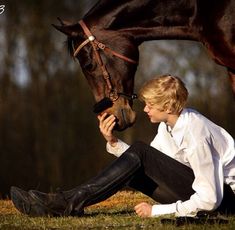 a young man sitting on the ground next to a brown horse with his head down