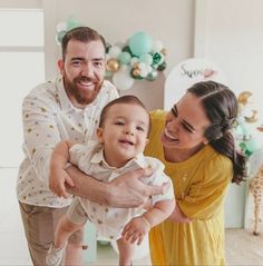 a man and woman holding a baby in front of a wall with balloons on it