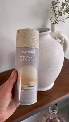 a person holding up a can of stone in front of a white vase on a shelf