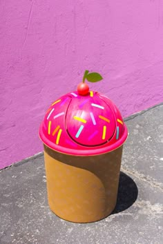 a pink cupcake with sprinkles and a cherry on top sits in front of a purple wall