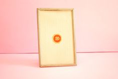 a white and gold frame with an orange circle on the front is against a pink background