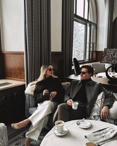 a man and woman sitting on a couch in a room with large windows, holding coffee cups