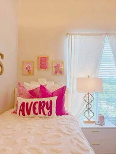 a white bed topped with pillows next to a window covered in pink and white curtains