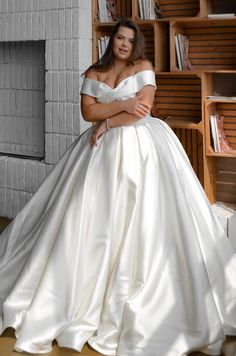 a woman in a white wedding dress leaning on a bookshelf with her arms crossed