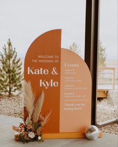 an orange and white wedding welcome sign with flowers on the ground in front of it