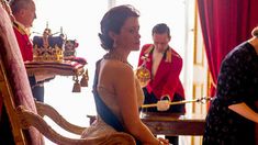 a woman in a dress sitting on a chair with other people standing around the room