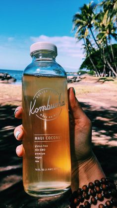 a person holding up a bottle of kombura on the beach with palm trees in the background