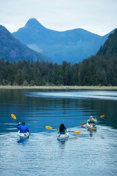 three people in kayaks paddling on the water with mountains in the background,