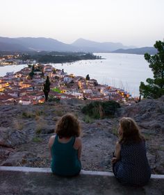 two women sitting on the edge of a cliff looking out over a town and water