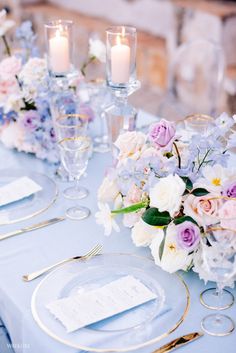 the table is set with white and purple flowers, silverware, and gold rimmed place settings