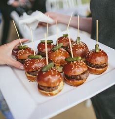 mini burgers with toothpicks are on a white platter while people stand in the background