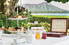 a table that has some food on it and drinks in front of the water with mountains in the background