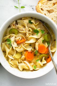 a white bowl filled with chicken noodle soup next to a slice of bread on a table