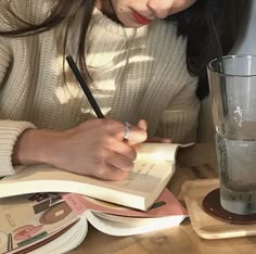 a woman sitting at a table writing on a book with a glass of water in front of her