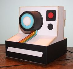 an origami polaroid camera sitting on top of a wooden table next to a red light