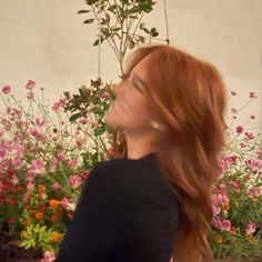 a woman with long red hair standing in front of some flowers and looking into the distance