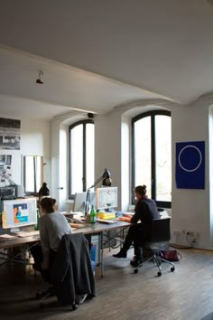 two people are sitting at a desk in an office with large windows looking out onto the street