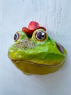 a green frog with a red hat on its head hanging from a white wooden wall