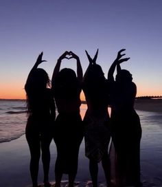 three girls standing in front of the ocean making a heart shape with their hands at sunset