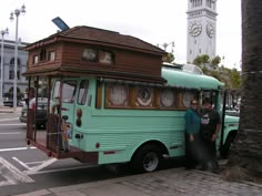 a man standing next to a green bus parked on the side of a road with a clock tower in the background