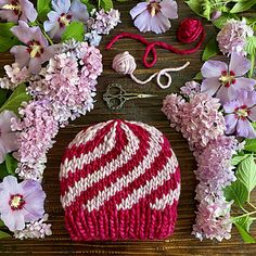 a red and white knitted hat next to pink flowers on a wooden table with scissors