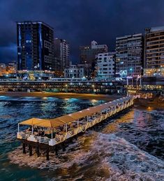 a pier in the middle of some water with buildings and lights on it at night