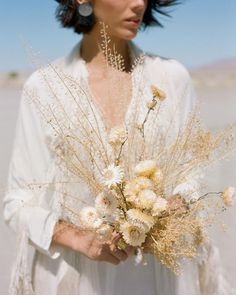 a woman in a white dress holding a bouquet of wildflowers and grasses with her hair blowing in the wind