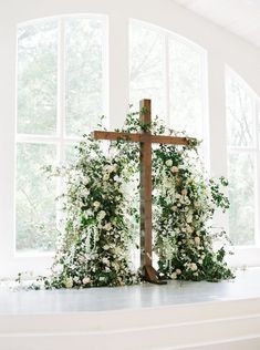 a wooden cross sitting in front of a window covered in white flowers and greenery