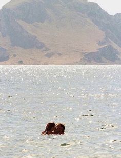 two people are swimming in the ocean with mountains in the background and one person is kissing the other