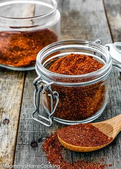 two jars filled with red chili seasoning on top of a wooden table next to a spoon