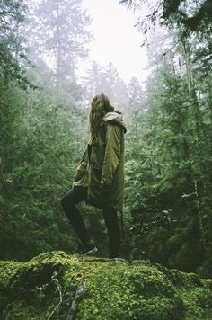 a woman standing on top of a moss covered rock in the woods with trees behind her